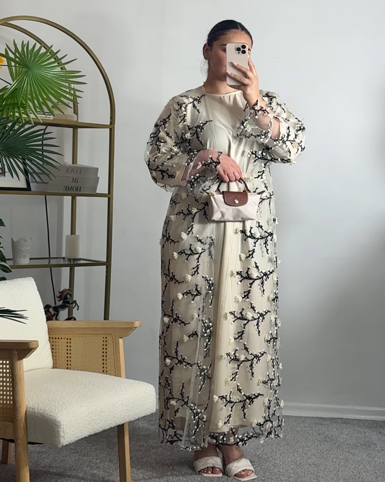Person wearing a floral dress taking a mirror selfie in a room with a plant and shelf.